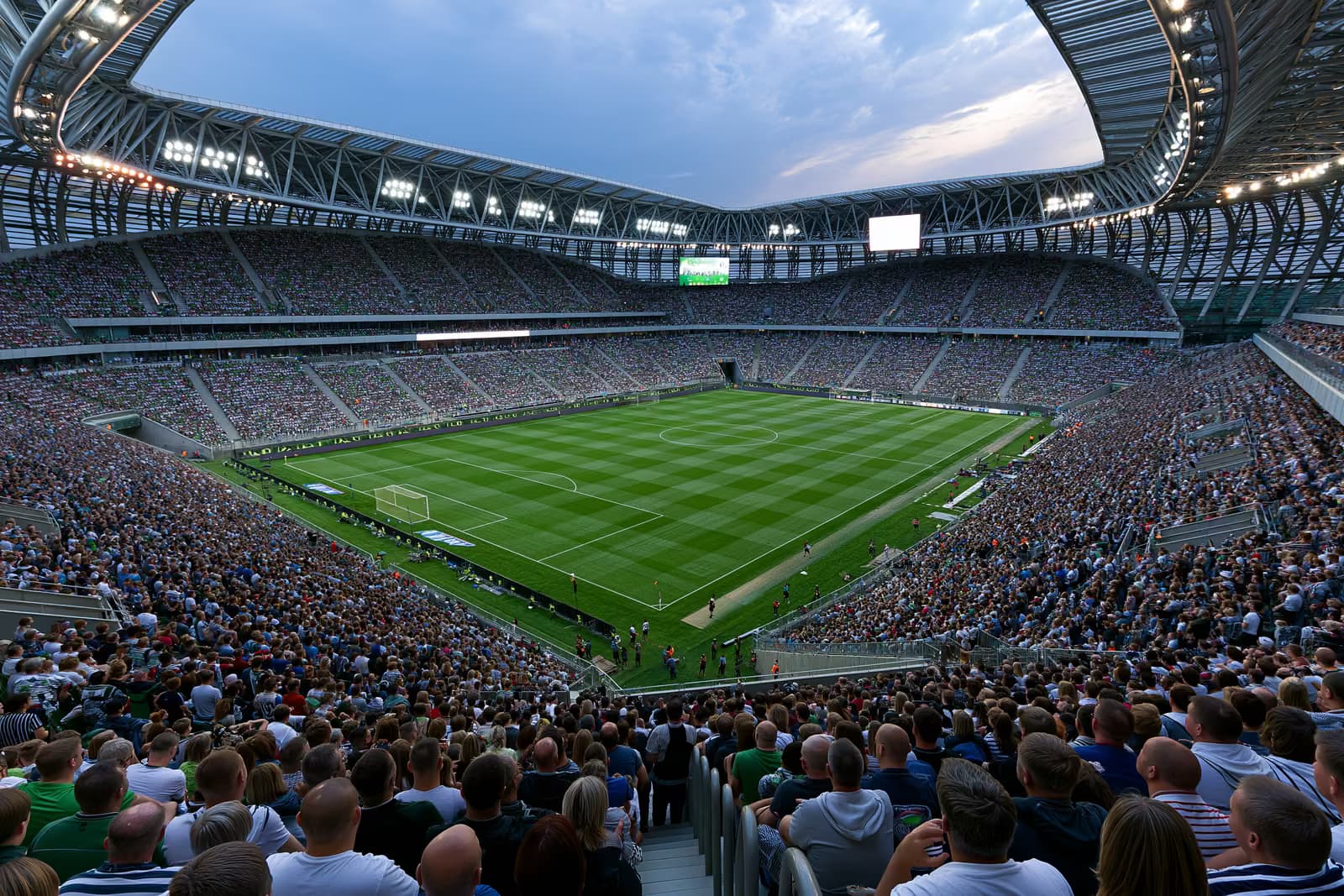 Egy hatalmas futballstadion, amely nyári estén reflektorok alatt teli szurkolókkal teli.