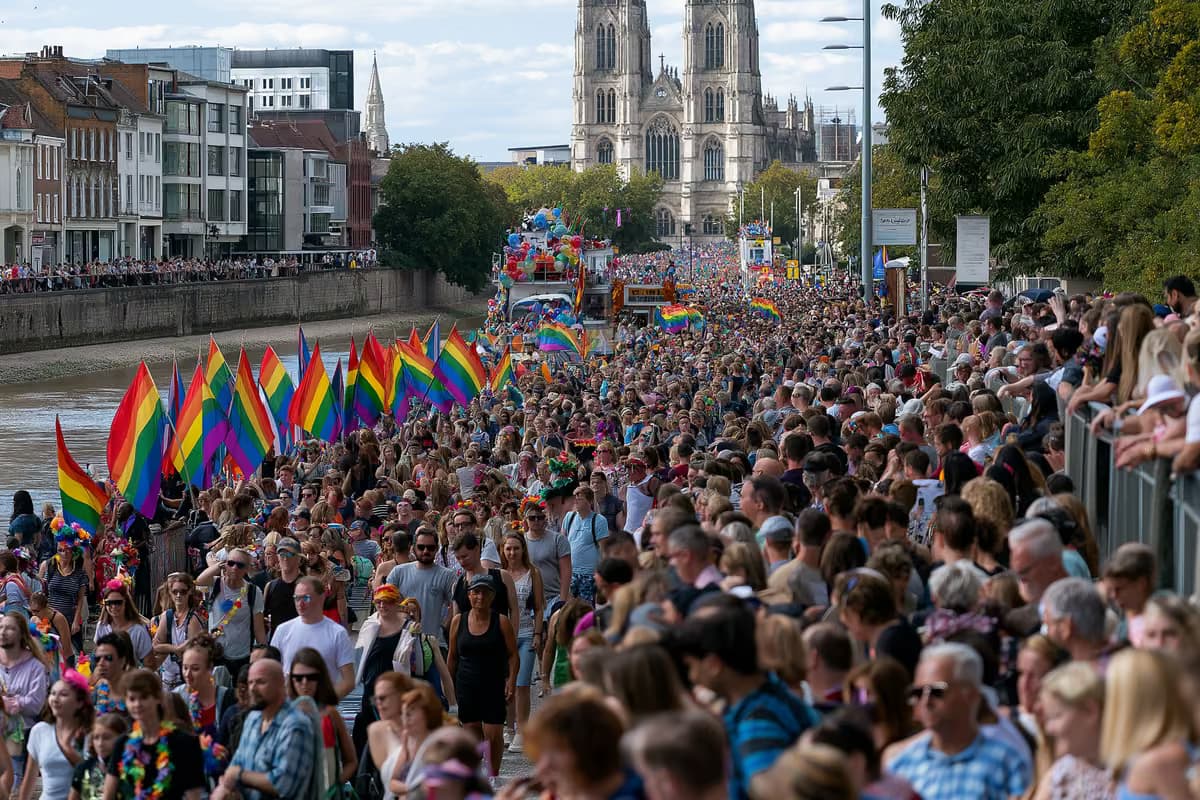 Szivárványos zászlók és feldíszített kocsik mozognak egy zsúfolt folyóparti utcán.