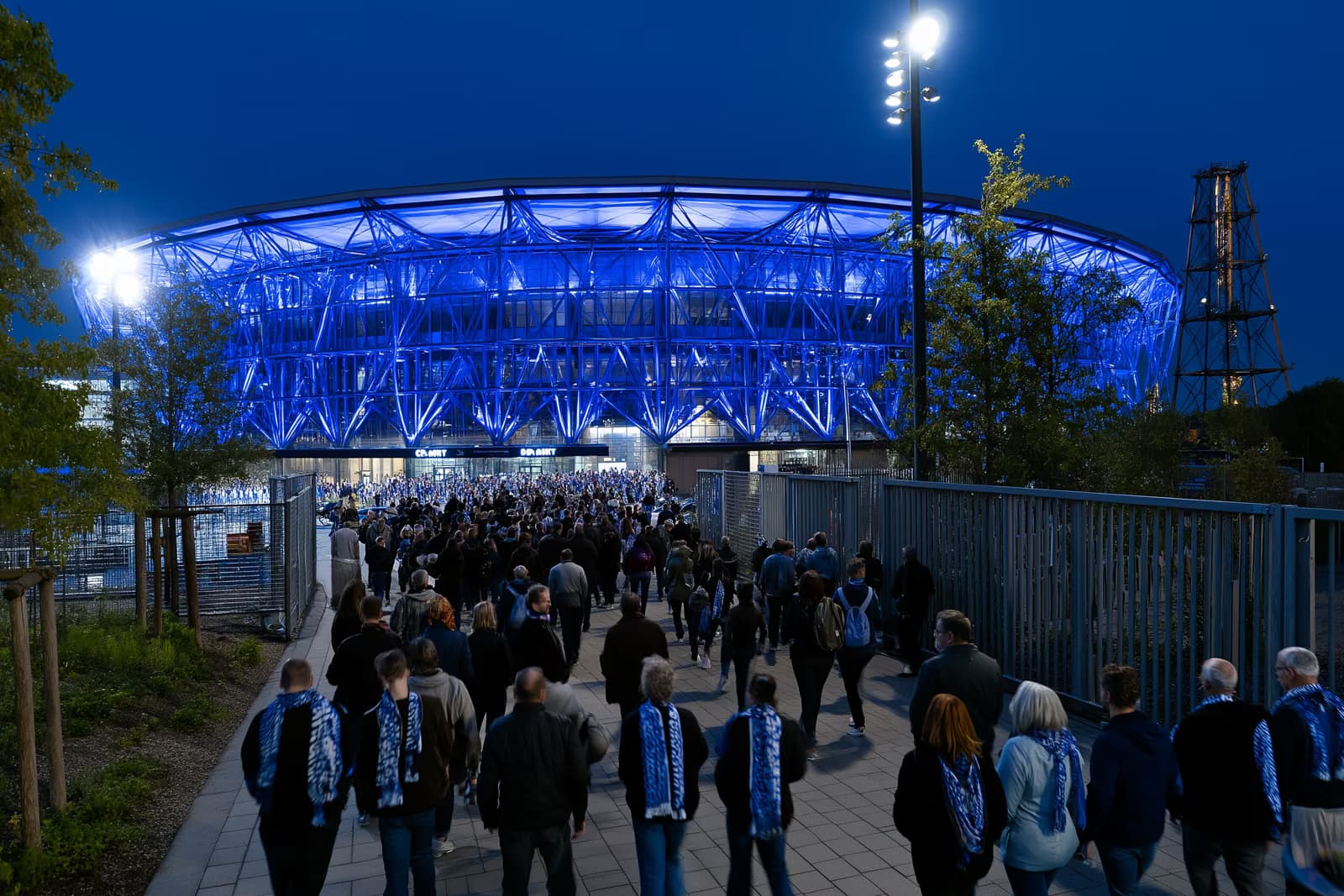 Egy Ruhr-vidéki futballstadion királykék fényben ragyog éjszaka, miközben a sálas szurkolók a kapu felé özönlenek a Gosens Schalke leigazolása után.